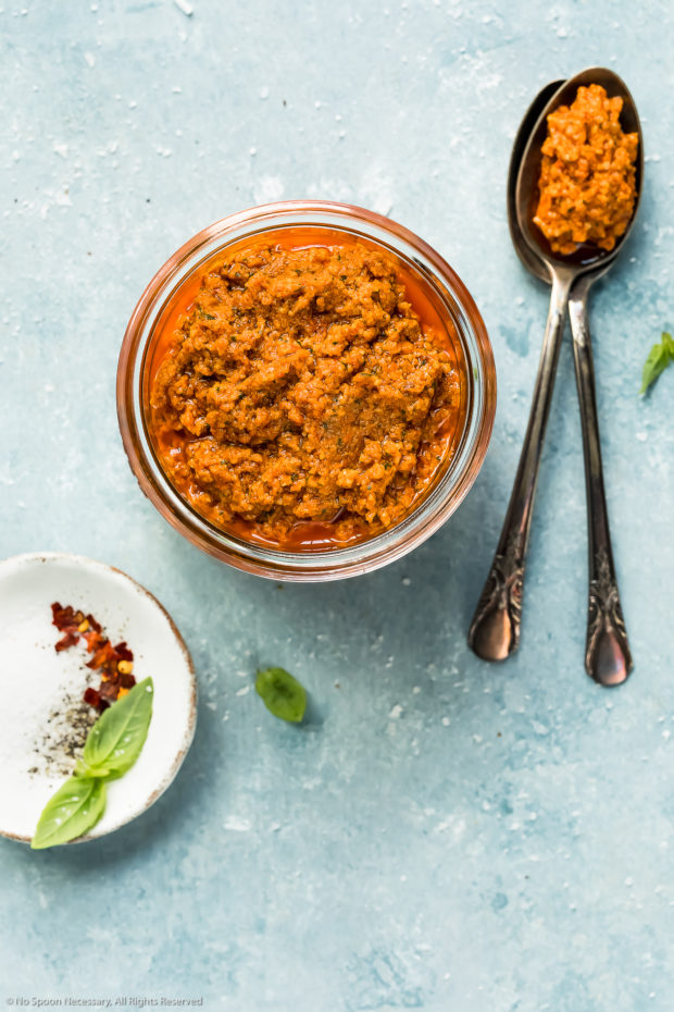 Overhead photo of Sun-Dried Tomato Pesto in a glass jar with a spoon of pesto and a ramekin of fresh basil leaves, salt, pepper and crushed red pepper flakes next to the jar.
