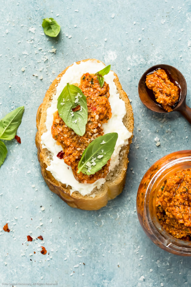 Overhead photo of a slice of toasted artisan bread topped with whipped cheese, pesto and basil leaves with a jar of pesto next to the bread - photo of how to use sun-dried tomato pesto.