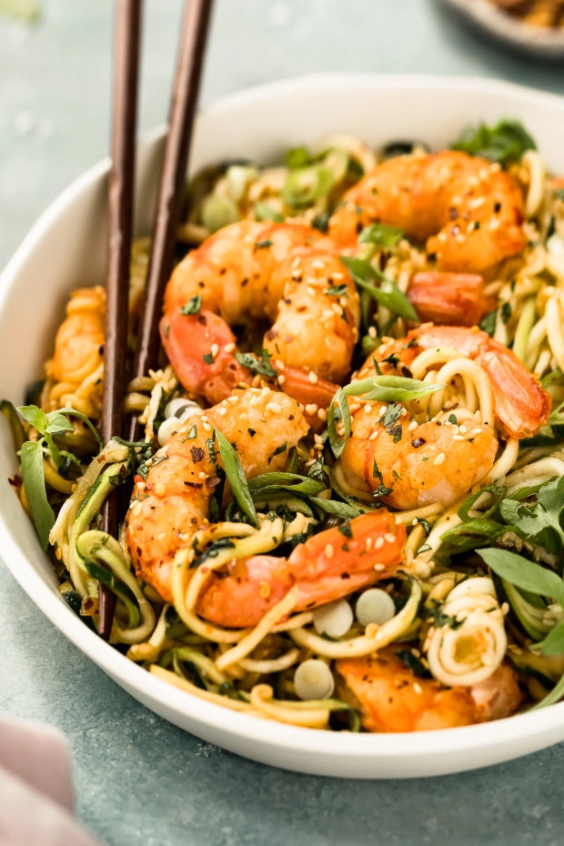 Angled photo of Vegetable Noodle Stir Fry topped with shrimp in a white bowl with a pair of chopsticks inserted into the noodles.