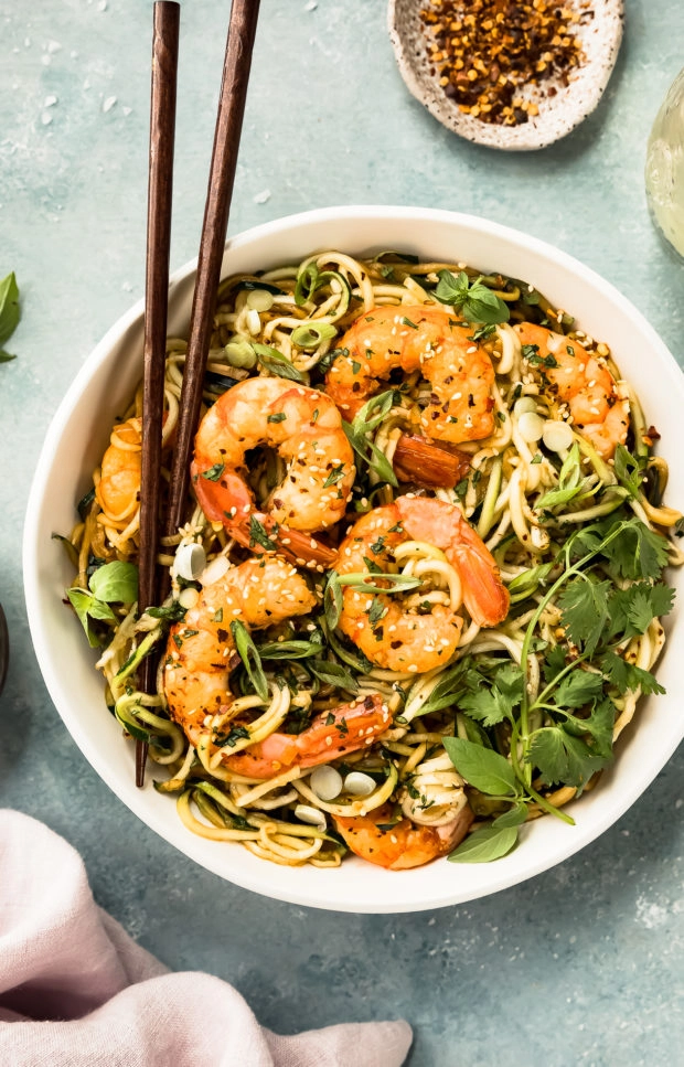 Overhead photo of Zucchini Vegetable Noodle Stir Fry topped with shrimp in a white bowl with a pair of chopsticks inserted into the noodles.