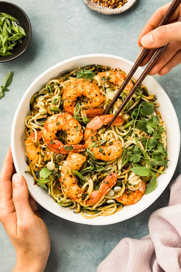 Overhead photo of Healthy Noodle Stir Fry topped with shrimp in a white bowl with a hand holding a pair of chopsticks and picking a shrimp up and out of the bowl.