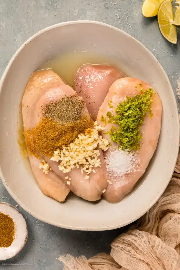 Overhead photo of four raw chicken breasts sprinkled with ground cumin, kosher salt, ground pepper, minced garlic and lime zest in a large mixing bowl.