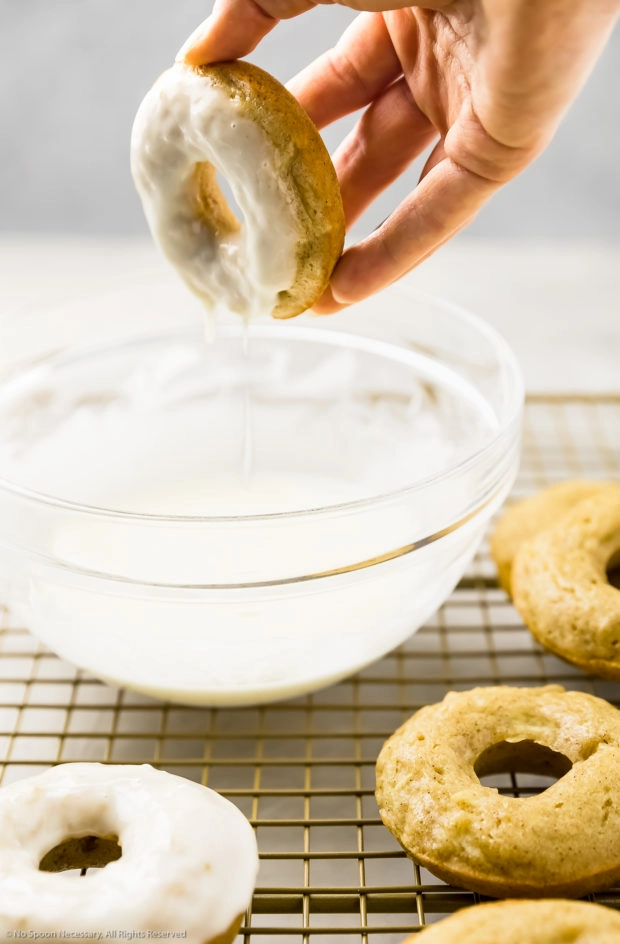 Angled photo of a hand dipping a cider donuts into a bowl of vanilla glaze with other donuts surrounding the bowl of glaze - photo of step 9 of the recipe.