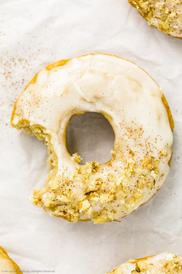 Overhead shot of a single glazed Cider Donut with a bite taken out of it on wrinkled parchment paper with cinnamon sprinkled on the donut and paper.