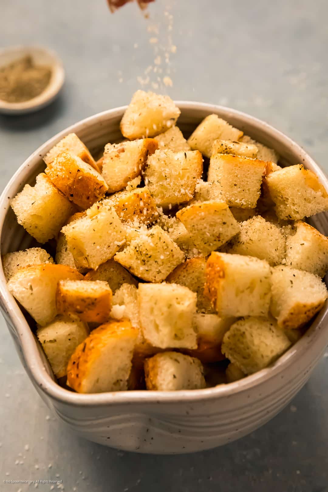 Action photo of a hand sprinkling freshly grated parmesan cheese on a bowl of bread croutons.