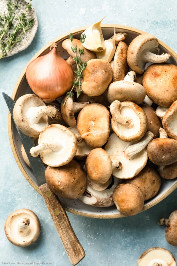 Overhead photo of assorted raw whole mushrooms in a neutral colored bowl with a knife and ramekin of fresh thyme next to the bowl - photo of the main ingredient in easy sauteed mushrooms recipe.