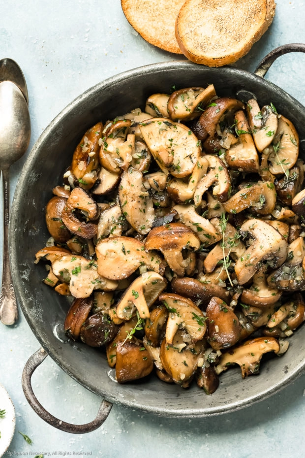 Overhead photo of sauteed mushrooms topped with thyme in a skillet with two pieces of toasted bread and spoons surrounding the skillet.