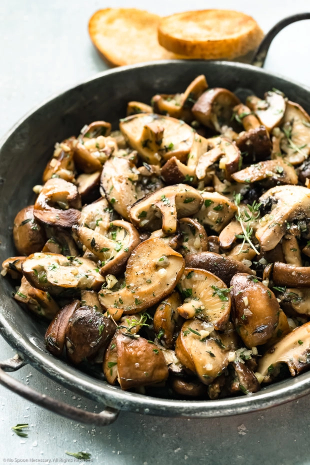 Angled photo of sauteed mushrooms topped with thyme in a skillet with two pieces of toasted bread blurred behind the skillet.