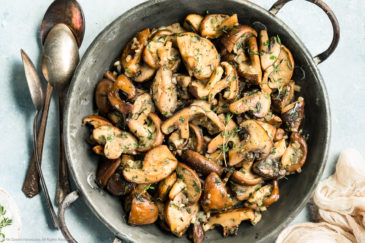 Overhead photo of sauteed mushrooms topped with thyme in a skillet with spoons and a neutral colored linen napkin surrounding the skillet.