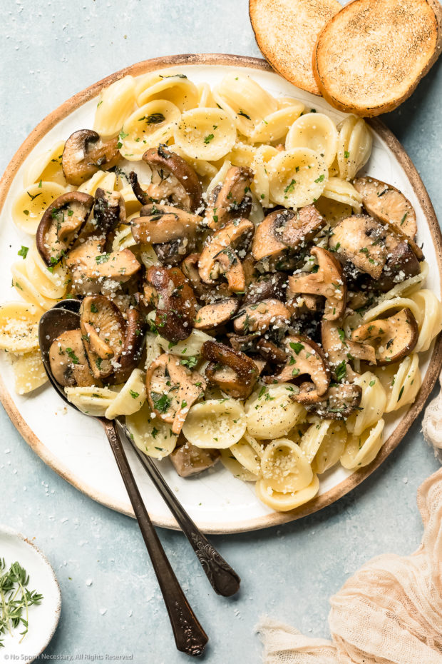 Overhead photo of pasta topped with butter sauteed mushrooms on a a white plate with serving spoons inserted into the pasta and two pieces of toasted bread, a neutral colored napkin and ramekin of fresh thyme next to the plate - photo of what to do with sauteed mushrooms.