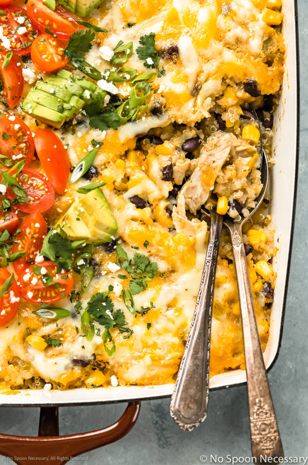 Overhead, close-up photo of a cheesy Mexican Chicken Bake with serving spoons inserted into the casserole showcasing the rotisserie chicken, quinoa and vegetable filling.
