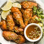 Overhead, landscape photo of Chinese Chicken Wings garnished with lime wedges and sesame seeds in a paper-lined serving bowl with a ramekin of Asian wing sauce next to the wings.