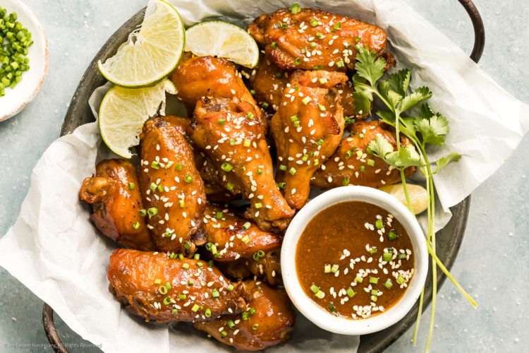 Overhead, landscape photo of Chinese Chicken Wings garnished with lime wedges and sesame seeds in a paper-lined serving bowl with a ramekin of Asian wing sauce next to the wings.