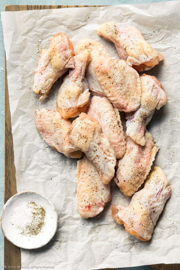 Overhead photo of raw chicken wings seasoned with salt and pepper on a parchment paper lined cutting board.