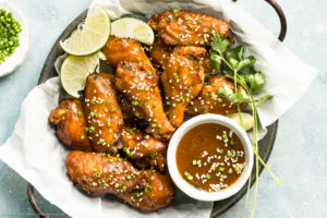 Overhead, landscape photo of Crockpot Asian Chicken Wings garnished with lime wedges and sesame seeds in a parchment paper lined serving bowl with a ramekin of Asian wing sauce next to the wings.