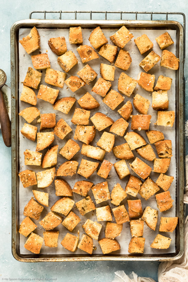 Overhead photo of seasoned and baked french bread cubes (croutons) spread out on a parchment paper lined sheet pan - photo of the step 5 of homemade crouton recipe.