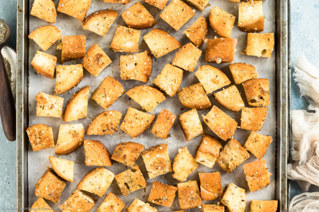 Overhead photo of homemade croutons on a parchment paper lined baking sheet with a ramekin of italian seasoning and neutral colored linen napkin next to the pan.
