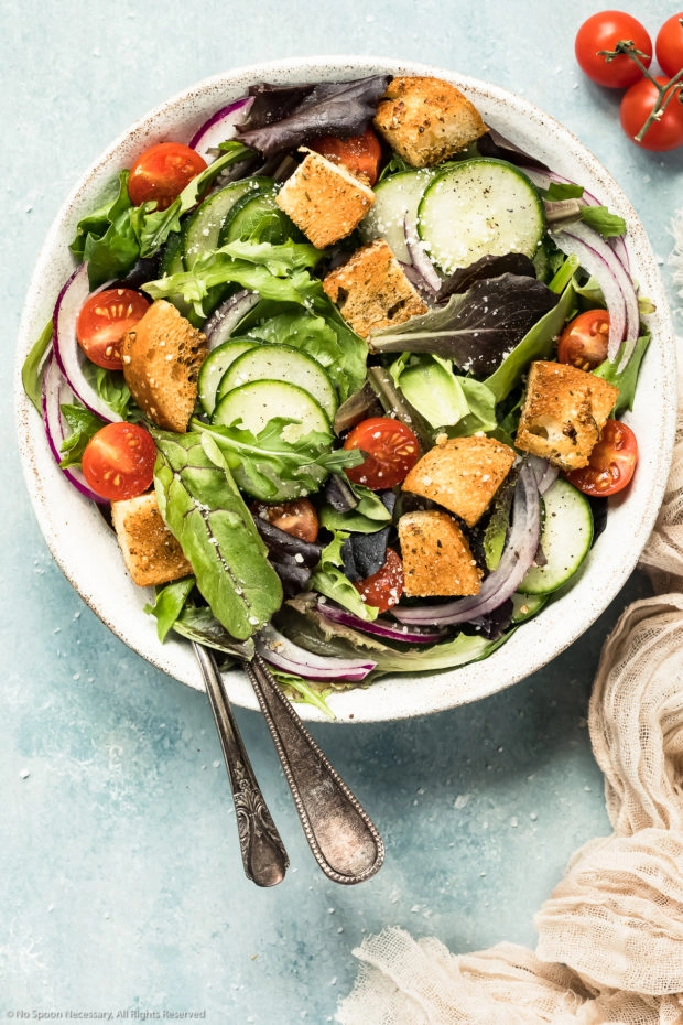 Overhead photo of a simple green salad topped with tomatoes, cucumbers, cucumbers and homemade croutons in a white bowl with a neutral colored linen napkin next to the bowl - photo of how to use easy homemade croutons.