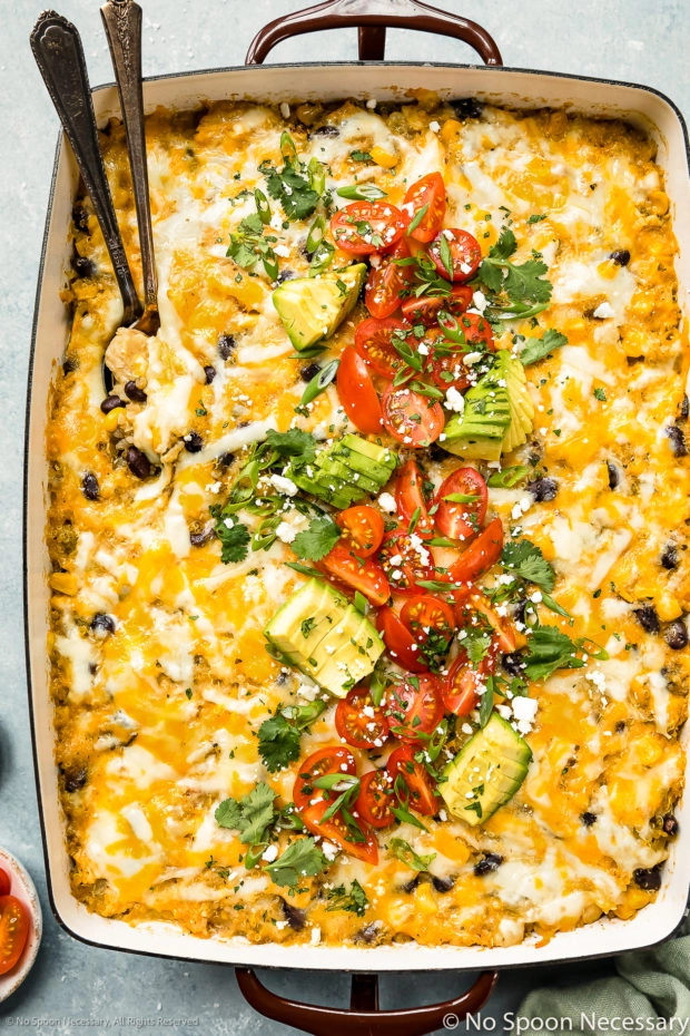 Overhead photo of cheesy Shredded Chicken Casserole garnished with diced tomatoes, sliced avocados and fresh cilantro with two serving spoons inserted into the dish.
