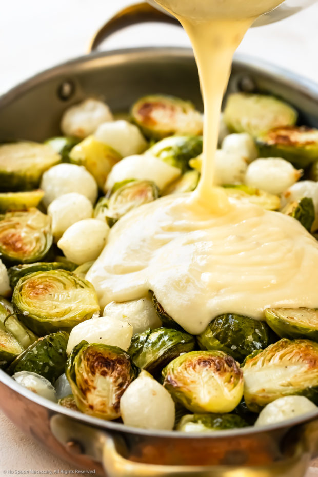 Angled photo of gruyere cheese sauce being poured over partially roasted brussels sprouts.
