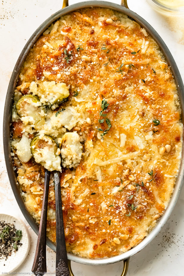 Overhead photo of Brussels Sprouts au Gratin in an oval baking dish with two spoons inserted into the casserole and a wine glass next to the dish.