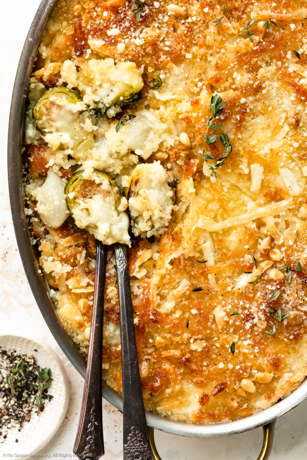Overhead, close up photo of brussels sprouts gratin casserole in an oval baking pan with a spoon inserted into the casserole dish exposing the white cheese sauce smothered brussels sprouts.