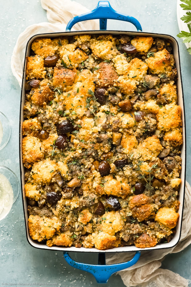 Overhead photo of Roasted Chestnut Stuffing made with cornbread and sausage in a large blue baking dish with glasses of wine and a beige napkin next to the dish.