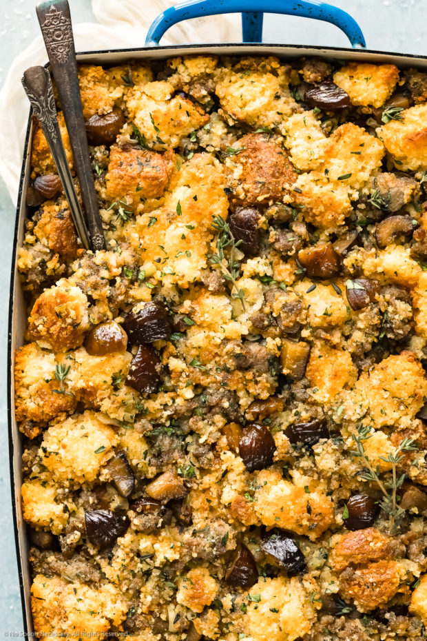 Overhead, close-up photo of Chestnut Stuffing made with sausage and cornbread in a blue baking dish with serving spoons inserted into the stuffing.