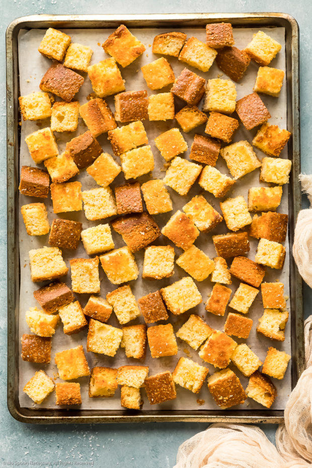 Overhead photo of cubed, toasted cornbread spread out on a parchment paper-lined baking sheet.
