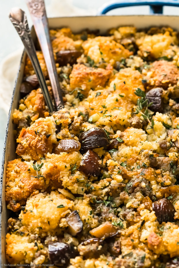 Angled, close-up photo of Thanksgiving Stuffing made with cornbread, chestnuts and sausage in a baking dish with serving spoons inserted into the stuffing.
