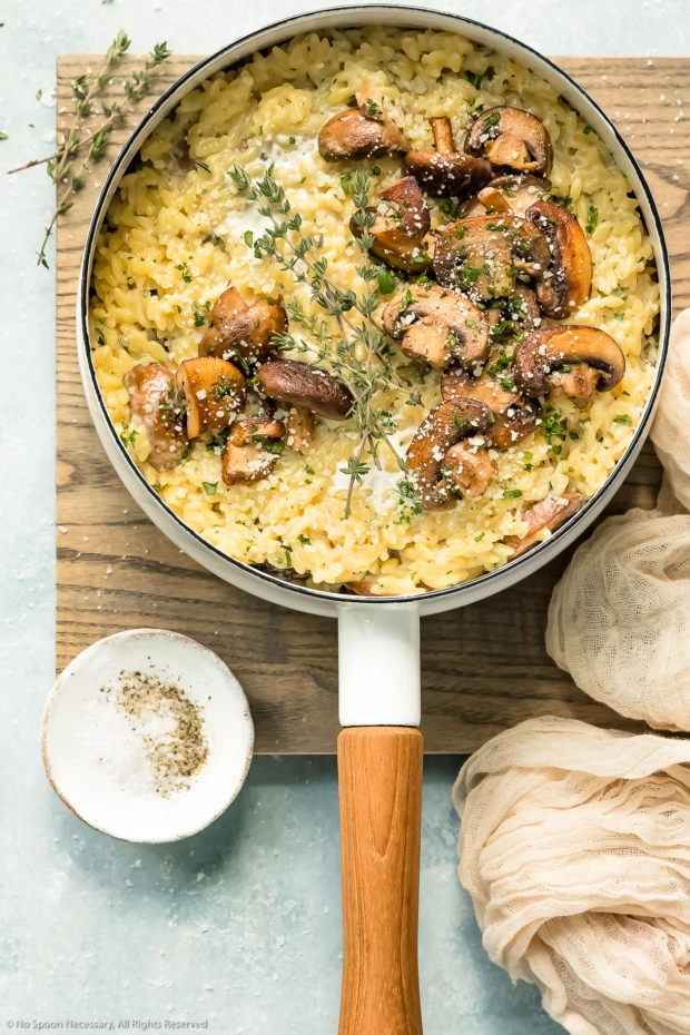 Overhead photo of Mushroom Orzo Risotto garnished with fresh thyme and grated parmesan in a white saucepan with a neutral colored linen and ramekin of salt and pepper next to the pan.
