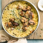 Overhead, landscape photo of Mushroom Orzo Risotto garnished with fresh thyme and grated parmesan in a white saucepan with a neutral colored linen and ramekin of salt and pepper next to the pan.