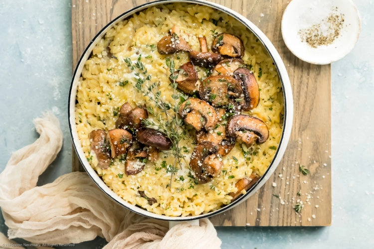 Overhead, landscape photo of Mushroom Orzo Risotto garnished with fresh thyme and grated parmesan in a white saucepan with a neutral colored linen and ramekin of salt and pepper next to the pan.