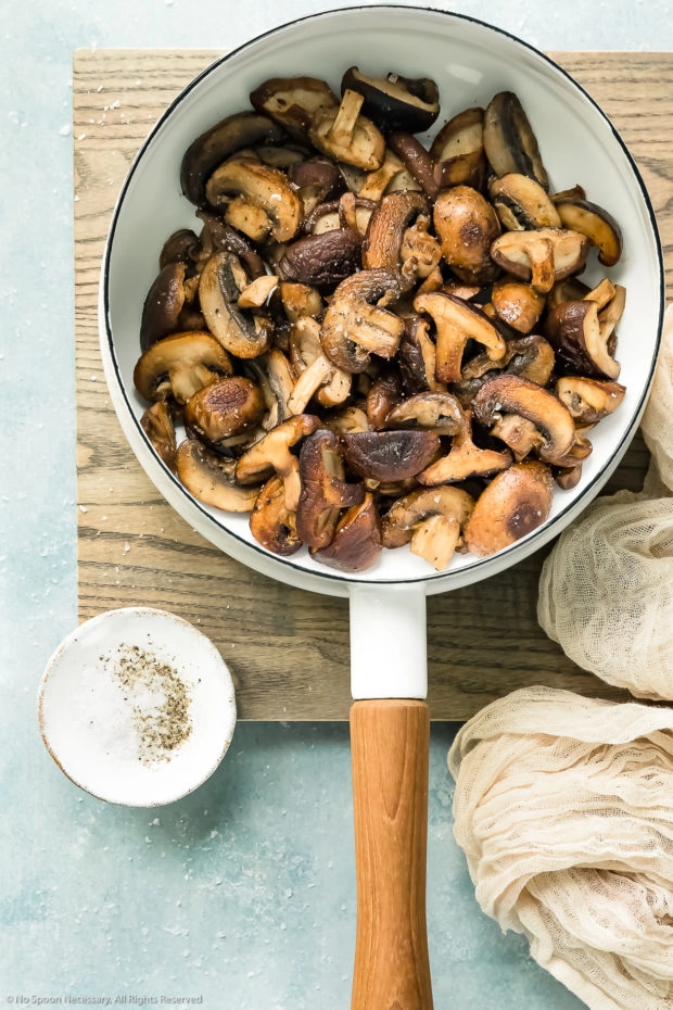 Overhead photo of sauteed mushrooms in a white saucepan on a wood surface with a neutral colored linen and ramekin of salt and pepper next to the pan - photo the first step of the recipe.