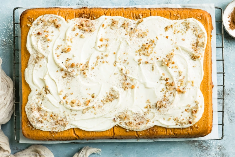 Overhead, landscape photo of Pumpkin Pie Cake topped with swirled maple cream cheese frosting on a wire rack with a ramekin of pumpkin pie spice and a neutral linen next to the cake.