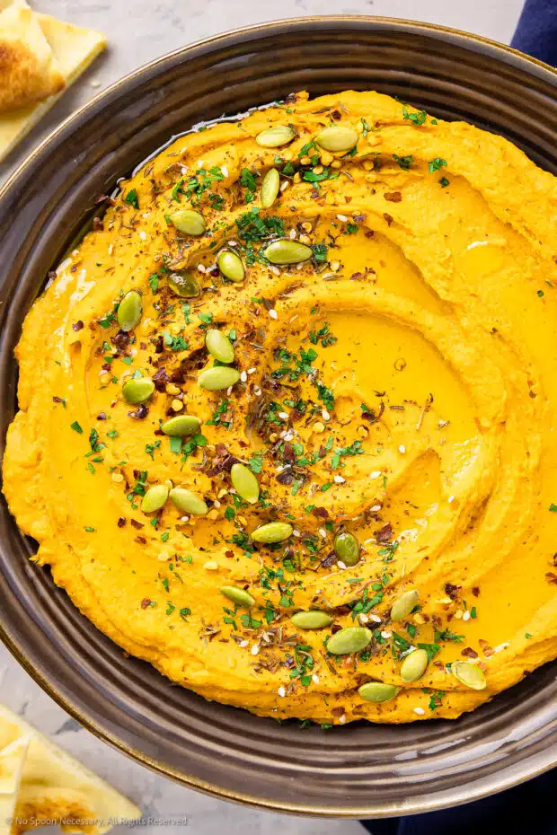Overhead photo of potato hummus garnished with herbs and nuts in a large serving bowl with pita chips next to the bowl.
