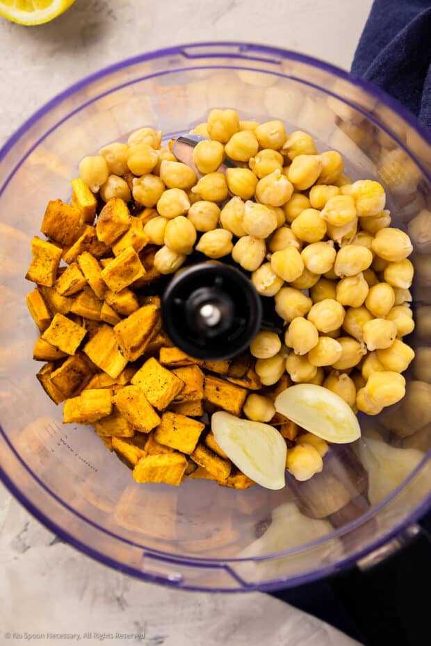 Overhead photo of chickpeas, diced roasted sweet potatoes and garlic cloves in the bowl of a food processor.