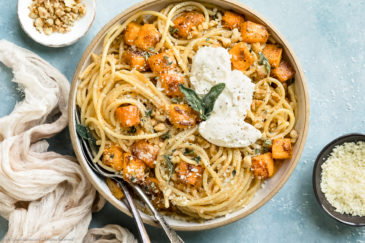 Overhead photo of Butternut Squash Pasta topped with whipped ricotta and crispy sage leaves in a white serving bowl with a tan napkin and ramekins of walnuts and grated parmesan off to the side of the bowl.