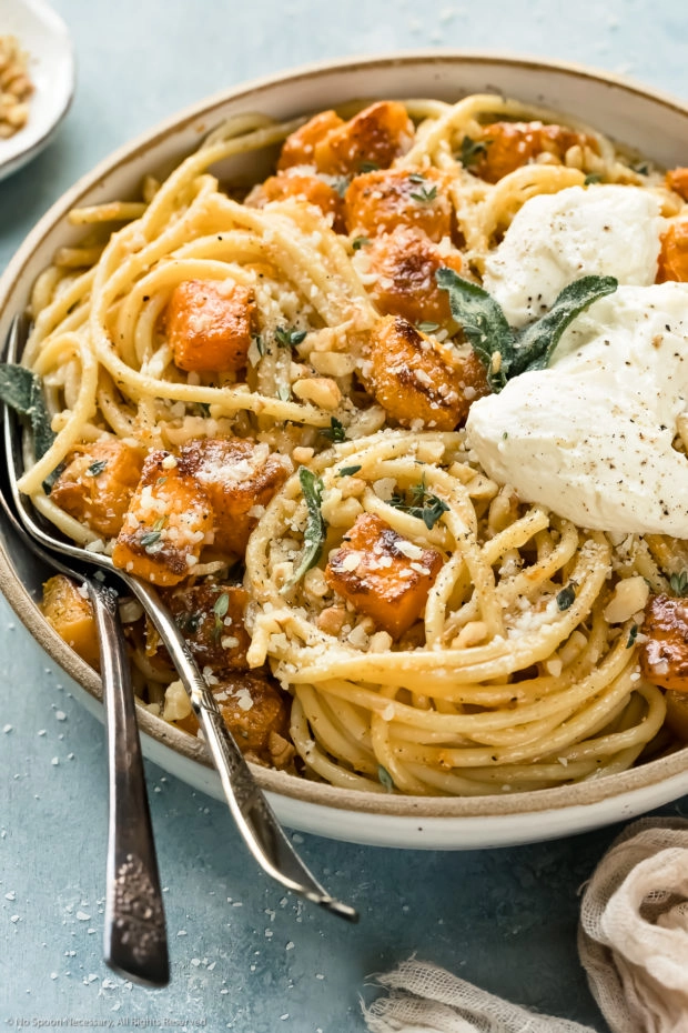 Angled photo of Butternut Squash Spaghetti Pasta topped with whipped ricotta, toasted walnuts and crispy sage leaves in a white serving bowl with serving spoons inserted into the pasta.