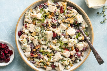 Overhead photo of Cranberry Chicken Salad garnished with sliced scallions and fresh thyme in a serving bowl with a spoon inserted into the salad and a ramekin of dried cranberries and a jar of dressing next to the bowl.