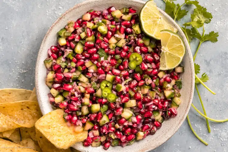 Overhead photo of cucumber salsa with jalapenos and pomegranate arils in a white bowl with tortilla chips on the side.