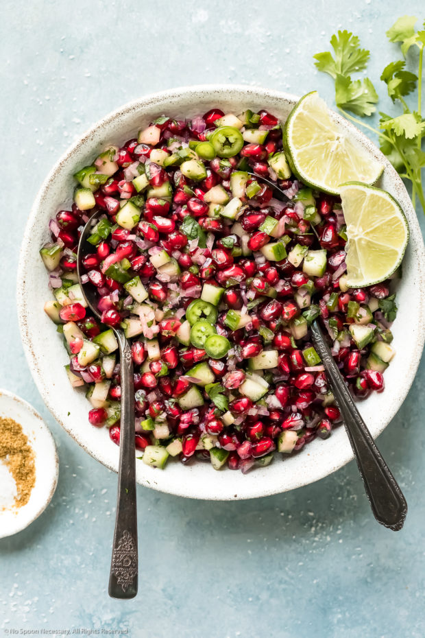 Overhead photo of Pomegranate Cucumber Salsa garnished with lime wedges in a white bowl with two serving spoons inserted into the salsa and fresh cilantro arranged next to the bowl.