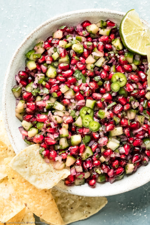 Overhead photo of Pomegranate Cucumber Salsa garnished with lime wedges in a white bowl with a tortilla chip sticking out of the salsa and more chips scattered next to the bowl.