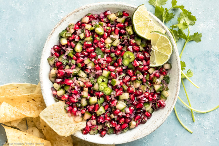 Overhead photo of Pomegranate Cucumber Salsa garnished with lime wedges in a white bowl with a tortilla chip sticking out of the salsa and more chips and fresh cilantro scattered next to the bowl.