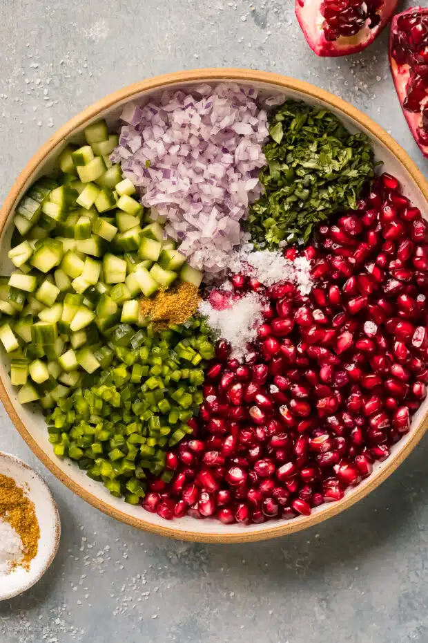 Overhead photo of pomegranate arils, diced cucumbers, diced jalapenos, diced red onion, chopped cilantro, and seasonings in a large salad bowl.