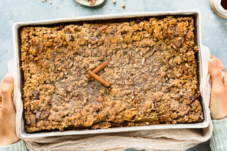 Overhead, landscape photo of two hands holding a Baked Cinnamon French Toast Casserole in a white baking dish with a jar of pure maple syrup and a ramekin of crushed pecans arranged around the baking dish.