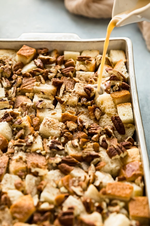 Angled photo of egg custard being poured over cubes of brioche layered with pecans and cinnamon in a white baking dish.