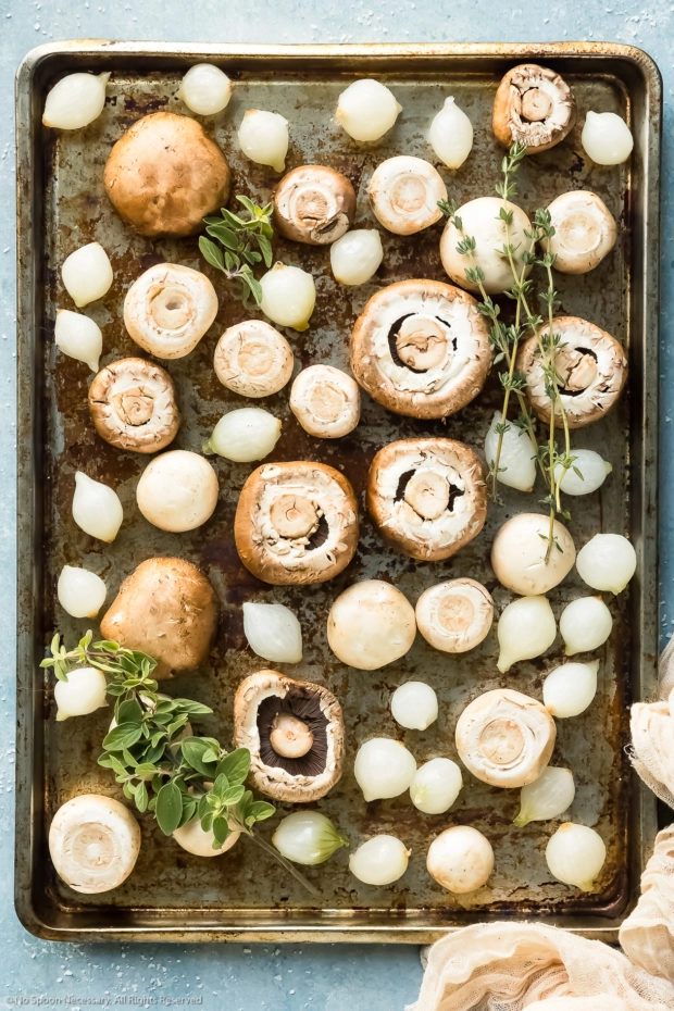 Overhead photo of fresh mushrooms and pearl onions topped with sprigs of fresh herbs on a rimmed baking sheet, before baking.