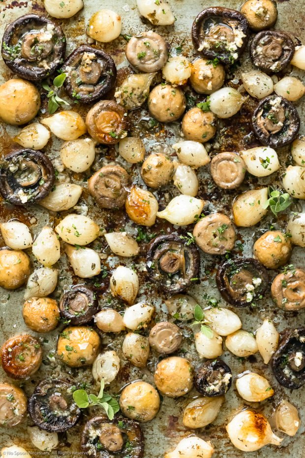 Overhead photo of roasted mushrooms and pearl onions topped with fresh herbs on a rimmed baking sheet, immediately after baking.