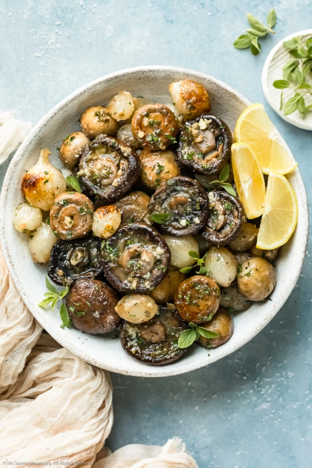 Overhead photo of Oven Roasted Mushrooms and Onions garnished with fresh herbs and lemon wedges in a white serving bowl with a pale tan napkin and ramekin of fresh oregano next to the bowl.